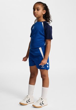 Ung pige står og har blå sportstøj på med hvide og marineblå accenter, hvide sokker og hvide sneakers med sorte chevron-logoer.