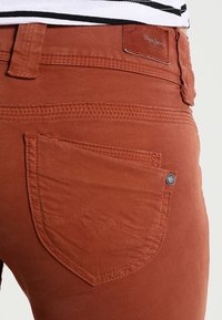 Ruskean väriset farkut, jotka on valmistettu puuvillasta ja joissa on tekstuuripinta. Takana on tasku, jossa on kontrastivärinen ompelu ja merkitty nahkapatch.