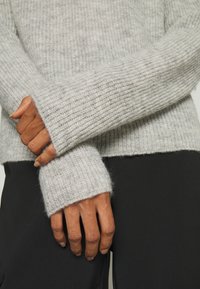 Mains croisées sur un pantalon noir, portant un pull en tricot côtelé gris clair à manches longues couvrant partiellement les mains.