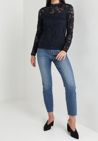 Marinblå spetsblus med lång ärm och blommönster, ihopparad med ljusblå jeans och svarta spetsiga klackar. Blusen har hög halsringning.