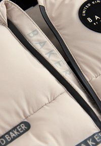 Vaate kevyessä beige sävyssä, jossa on mustat vetoketjut ja yksityiskohtia, pyöreä logo-ommel mustavalkoisena sekä harmaata painettua tekstiä.