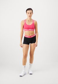 Rozā sporta krūšturis ar baltu "NIKE PRO" jostu, apvienots ar melniem šortiem, kuriem ir atbilstoša josta. Modelis valkā baltas sporta kurpes un zeķes.