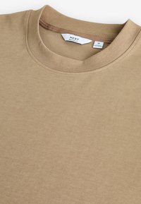 Beige T-Shirt mit Rundhalsausschnitt aus weichem Stoff. Bietet einen lässigen Schnitt, eine gerippte Kragenpartie und ein Etikett, das im Ausschnitt sichtbar ist. Breite, flache Textur.