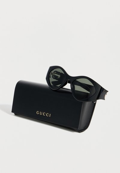 Μαύρα μεγέθη γυαλιά ηλίου ακουμπισμένα πάνω σε μαύρη θήκη Gucci με χρυσό λογότυπο, σε λευκό φόντο.