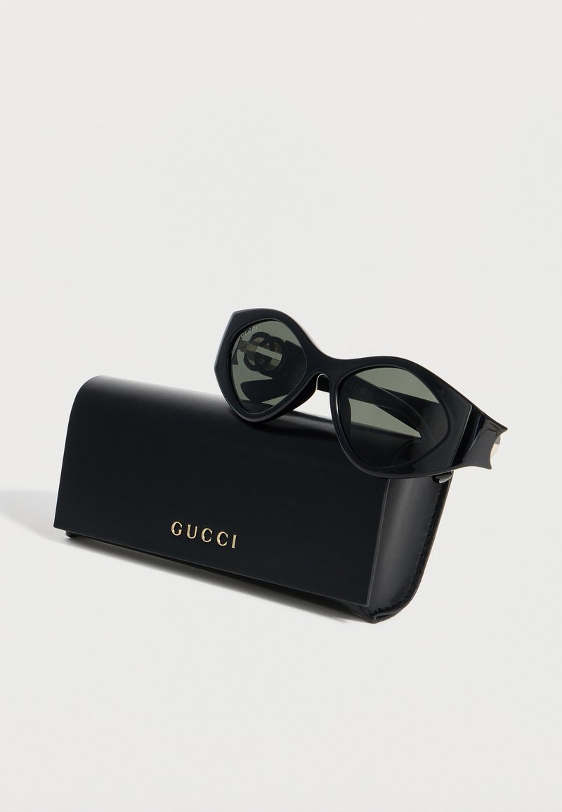 Schwarze übergroße Sonnenbrille, die auf einem schwarzen Gucci-Etui mit goldenem Logo liegt, vor einem schlichten weißen Hintergrund.
