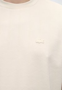 Κοντινή λήψη μπεζ υφασμάτινου πουκαμίσου με μικρό κεντητό λογότυπο κροκόδειλου στο στήθος, με στρογγυλή λαιμόκοψη.