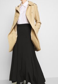 Gabardina beige sobre una camisa blanca, combinada con una falda negra fluida. La gabardina tiene botones de doble fila y un diseño clásico.