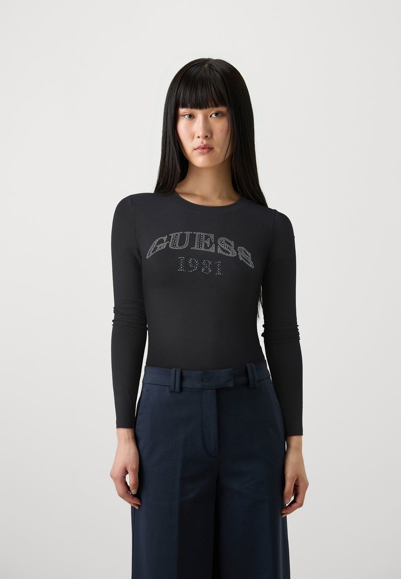 Maglietta nera a maniche lunghe con logo in strass "GUESS 1981". Materiale morbido, design aderente, abbinata a pantaloni navy a vita alta.