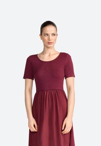 Robe bordeaux avec un corsage côtelé, des manches courtes et une jupe fluide. Détail logo brodé sur le devant. Texture lisse.