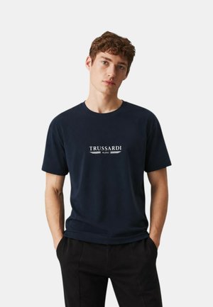 Giovane uomo con capelli ricci che indossa una t-shirt blu navy Trussardi Milano e pantaloni neri, in piedi con le mani in tasca su uno sfondo semplice.