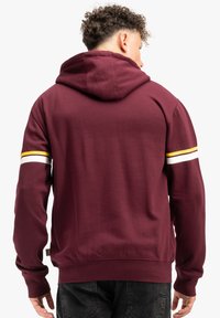 Bordová mikina s kapucňou na sťahovanie, rebrovanými manžetami a pásom. Obsahuje dve horizontálne pruhy v žltej a bielej farbe na rukávoch.