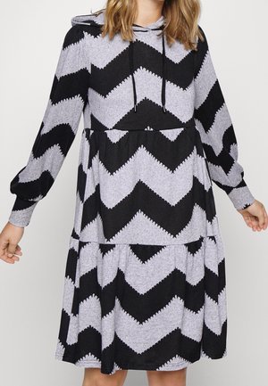 Femme portant une robe à capuche noire et grise à manches longues avec un motif zigzag et une jupe à volants, debout les bras légèrement étendus.