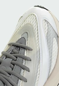 Harmaa ja valkoinen urheilukenkä, jossa on teksturoitu verkko-yläosa, näkyvät nauhat ja sileät yksityiskohdat, joiden muotoilu on kaareva ja kumipohja.