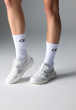 Legs wearing white athletic socks with black infinity logo and white mesh sneakers with textured soles on a gray background.