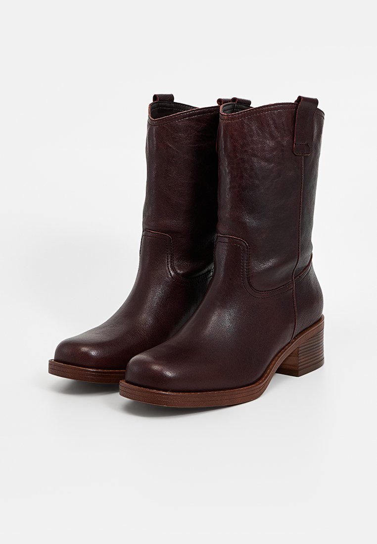Botas de tobillo de cuero marrón con una textura suave, puntera redondeada y tacón bajo apilado. Presentan costuras decorativas y lengüetas en la parte superior para facilitar su uso.