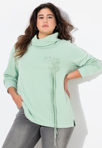 Top verde chiaro a maniche lunghe con collo a barchetta, design ricamato sul petto e lunghi nastri davanti. Tessuto morbido.