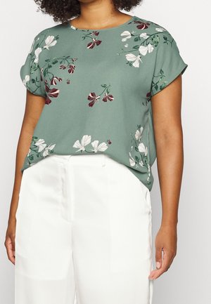 Vrouw die een groene bloemige blouse met korte mouwen draagt, gestoken in een witte broek met hoge taille, afgebeeld van nek tot heupen.