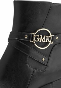 Bota corta de cuero negra con una hebilla circular en tono dorado que presenta las iniciales "GMK", con costuras mínimas y un acabado texturizado.