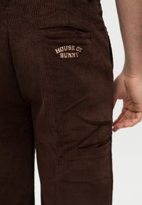 Κοντινό πλάνο καφέ παντελονιού από κοτλέ με τη ραφή "HOUSE OF SUNNY" πάνω από την δεξιά πίσω τσέπη και ένα χέρι που ακουμπά κοντά.