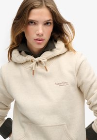 Felpa beige con cappuccio, dotata di cordini e dettagli marroni. Presenta una tasca frontale e il logo ricamato "SuperDry" sul petto.