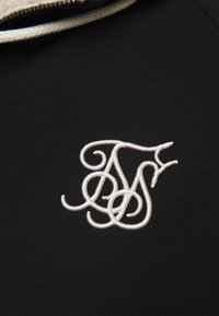 Tessuto nero con un primo piano di iniziali bianche ricamate. Il design presenta lettere eleganti e intrecciate con una finitura strutturata.