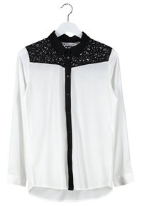 Chemise blanche à manches longues avec un panneau en dentelle noire sur le yoke. Dotée d'une patte de boutonnage à l'avant et de boutons noirs contrastants. Texture de tissu lisse.