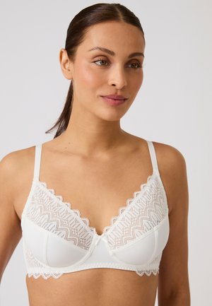 Donna con capelli castani legati indietro, che indossa un reggiseno di pizzo bianco con bordi smerlati, guarda leggermente verso destra su uno sfondo neutro.