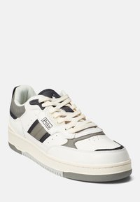 Zapatilla deportiva blanca con acentos grises, con una parte superior de cuero suave, puntera redonda, cuello acolchado y cordones planos. Diseño clásico de tres rayas.