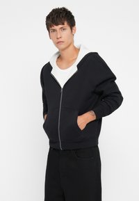 Casaco preto com fecho éclair e forro de lã branca, com bolsos tipo canguru e punhos canelados, apresentado por um modelo em fundo branco simples.