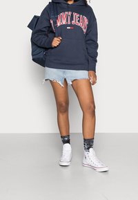 Námořnická modrá mikina s logem "TOMMY JEANS", otrhaná džínsová mini sukně, bílé vysoké tenisky a vzorované černé ponožky.