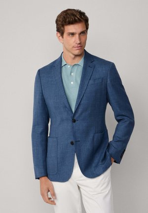 Blazer in Blau mit strukturiertem Stoff, Schößchen, zwei Vorderaschen und Ein-Knopf-Verschluss, kombiniert mit einem hellblauen Poloshirt und weißen Hosen.