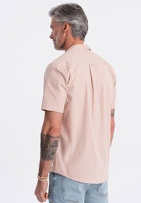 Pinken shirt met korte mouwen, een rugplooi, kraag en een zachte textuur; gedragen met lichtblauwe jeans. Kenmerkt een zichtbaar getatoeëerde arm.