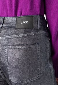 Tmavé denímové džínsy s lesklým povrchom, s čiernou visačkou s nápisom "JJXX" a štandardnými zadnými vreckami. Fialový pletený top je čiastočne viditeľný.