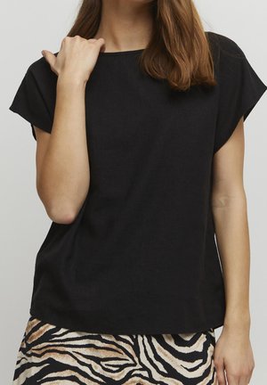 Kobieta w luźnej, czarnej bluzce z krótkim rękawem i spódnicy w paski zebry, z jedną ręką przy ramieniu, a drugą swobodnie ułożoną wzdłuż ciała.