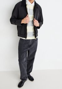 En person iført en sort zipjakke over en creme- og gråstribet polo skjorte, løse sorte jeans og sorte sneakers mod en hvid baggrund.