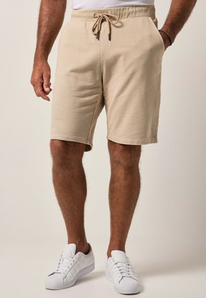 Beige knælange shorts med snøre, båret af en person med hvide sneakers og et armbånd på højre håndled.