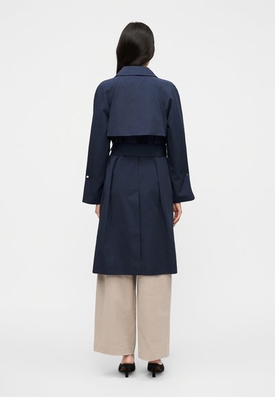 Ναυτικό παλτό τύπου τρανς με ζώνη στη μέση, φαρδιά μανίκια και κοντή λεπτομέρεια στο πίσω μέρος. Το μοντέλο φοράει ανοιχτό μπεζ παντελόνι με φαρδιά μπατζάκια και τακούνια.