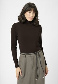 Mujer con cabello oscuro hasta los hombros, vistiendo un suéter de cuello alto marrón oscuro y una falda de cuadros de cintura alta con un delgado cinturón negro.