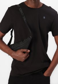 Camiseta negra de algodón con escote redondo y pequeño logotipo. La modelo sostiene un bolso bandolera negro texturizado con cremallera y correa ajustable.