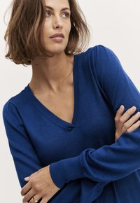 Modra v-necka pulover z dolgimi rokavi, izdelan iz teksturiranega pletenja. Rokavi so sproščenega kroja in vsebujejo rahlo gubanjem na ramenih.