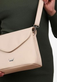 Beige kožená taška přes rameno s klopou, stříbrným kováním a embosovaným logem na přední straně. Viditelný nastavitelní popruh.