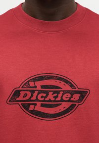 Κοντινό πλάνο ενός ατόμου που φοράει κόκκινη μπλούζα με στρογγυλή λαιμόκοψη και ξεθωριασμένο μαύρο λογότυπο Dickies στο κέντρο του στήθους.