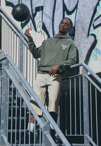 Oliven grøn hoodie med en tyr grafik, lyseblå denim shorts, sorte sneakers og en basketball, poseret på en metaltrappe med graffiti baggrund.