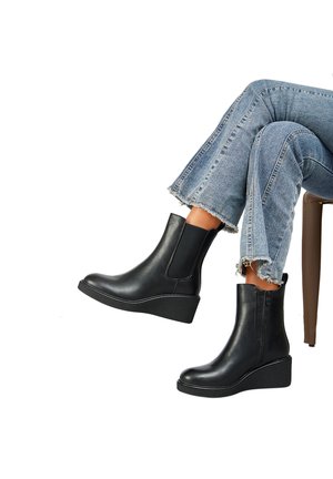 Personne portant des bottines compensées noires et un jean bleu à ourlet effiloché, assise jambes croisées sur fond blanc.