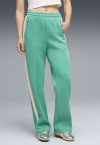 Γυναίκα με πράσινο αθλητικό παντελόνι με λευκές ρίγες στο πλάι και ασημένια αθλητικά παπούτσια, με το χέρι στην τσέπη, στέκεται μπροστά σε γκρι φόντο.