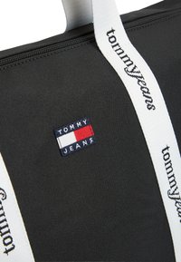 Μαύρη υφασμάτινη τσάντα ώμου με φερμουάρ, που διαθέτει λευκό λουρί με την εγγραφή "tommy jeans" και ένα μικρό λογότυπο σε μπλε ναυτικό, κόκκινο και λευκό.