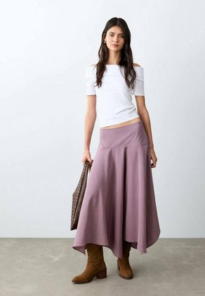 Frau trägt weiße schulterfreie Bluse, fliederfarbigen asymmetrischen Rock, braune Wildlederstiefel und hält eine braune, mit Nieten besetzte Handtasche vor schlichtem Hintergrund.