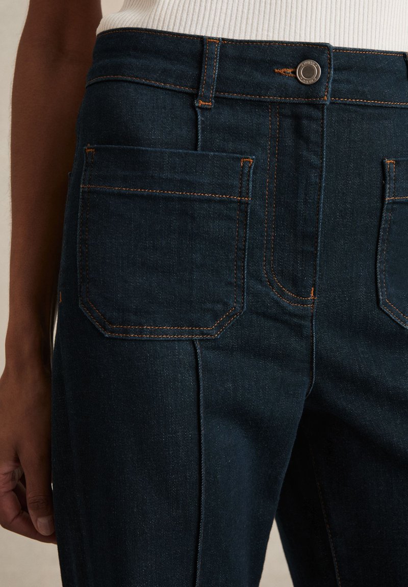 Mørkeblå denimjeans med markante forlommer og kontrastfuld orange syning, båret med en hvid ribbet top.