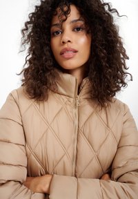 Beżowa pikowana kurtka puchowa z wysokim kołnierzem, z diamentowym przeszyciem, zapinana na zamek błyskawiczny w złotym odcieniu. Miękka faktura, lekka konstrukcja.