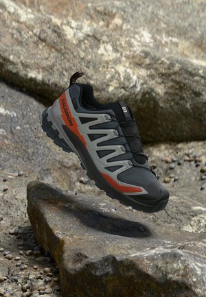 Chaussure de trail de couleur grise et orange flottant au-dessus d'un terrain rocheux.